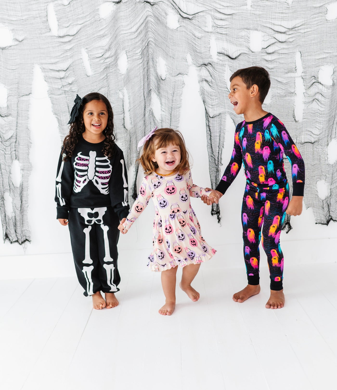 Three children in Halloween-themed outfits standing in front of a decorative white wall with black cut-out shapes.