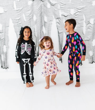Three children in Halloween-themed outfits standing in front of a decorative white wall with black cut-out shapes.