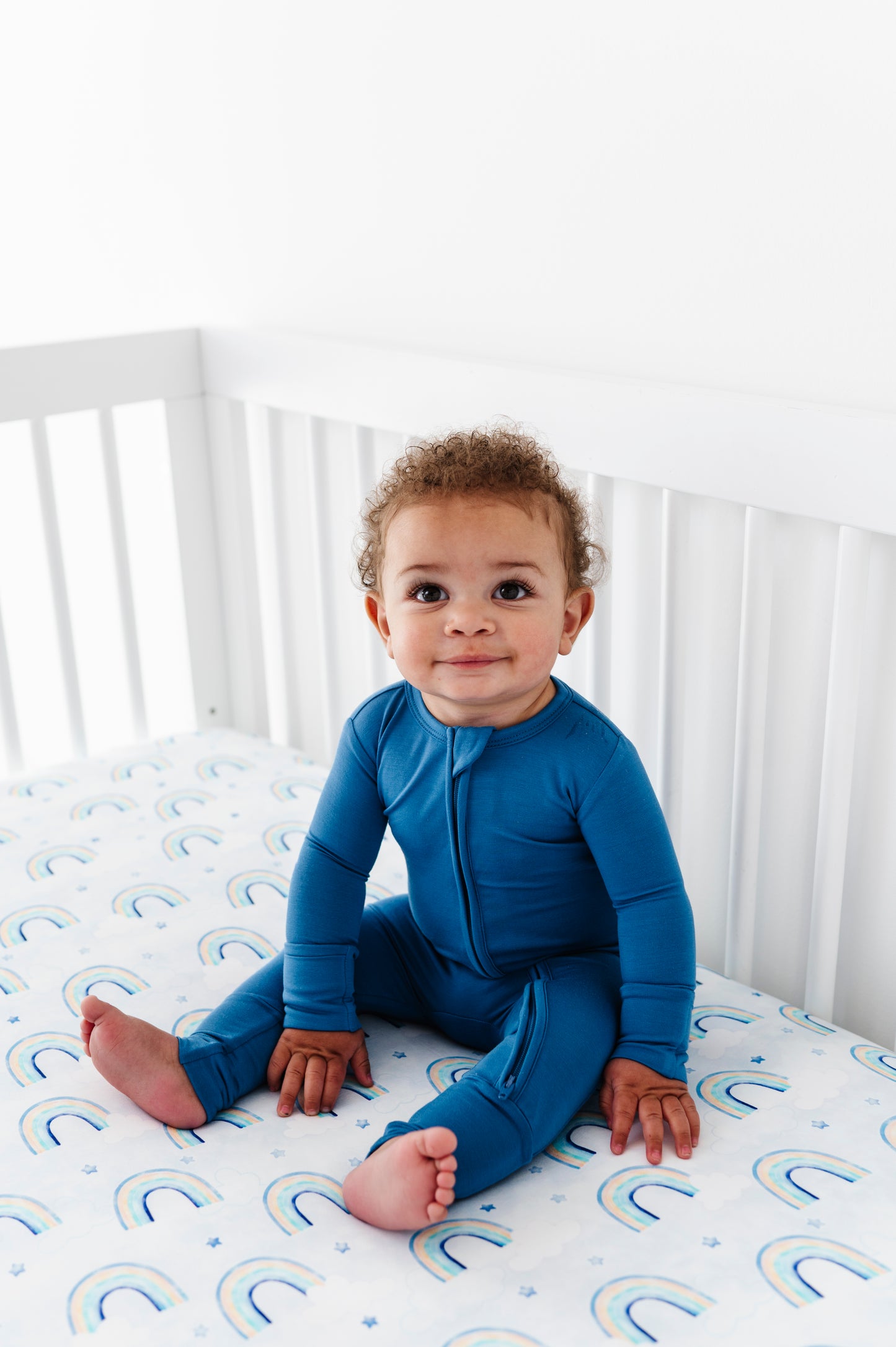 Child wearing a blue bamboo zip romper sitting on a crib with a white mattress featuring watercolor rainbow patterns.