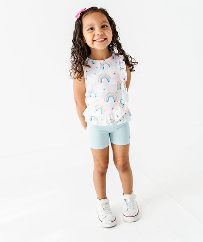 Young girl wearing a colorful rainbow bamboo cotton peplum top and light blue bamboo biker shorts on a white background