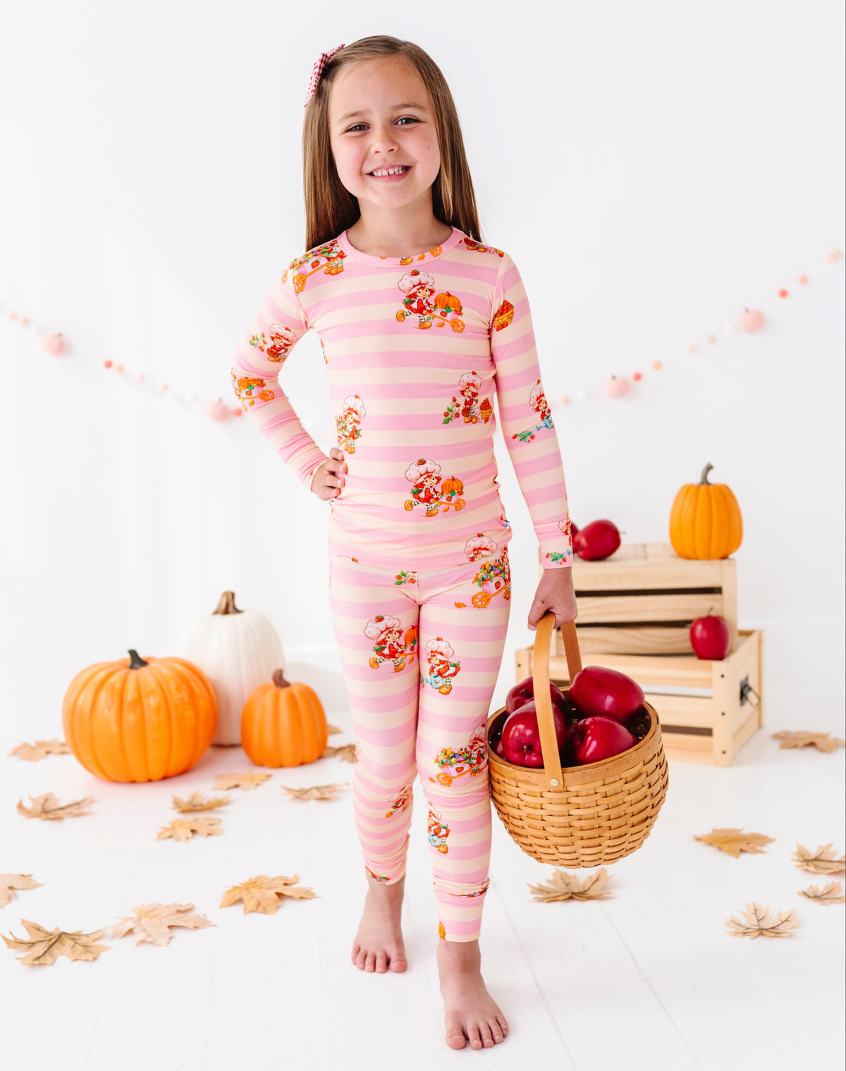 A young girl stands barefoot in a studio setting wearing a pink and white striped long-sleeve pajama set with a small floral print. She is holding a wicker basket filled with red apples, with pumpkins, wooden crates, and scattered autumn leaves on the floor around her.