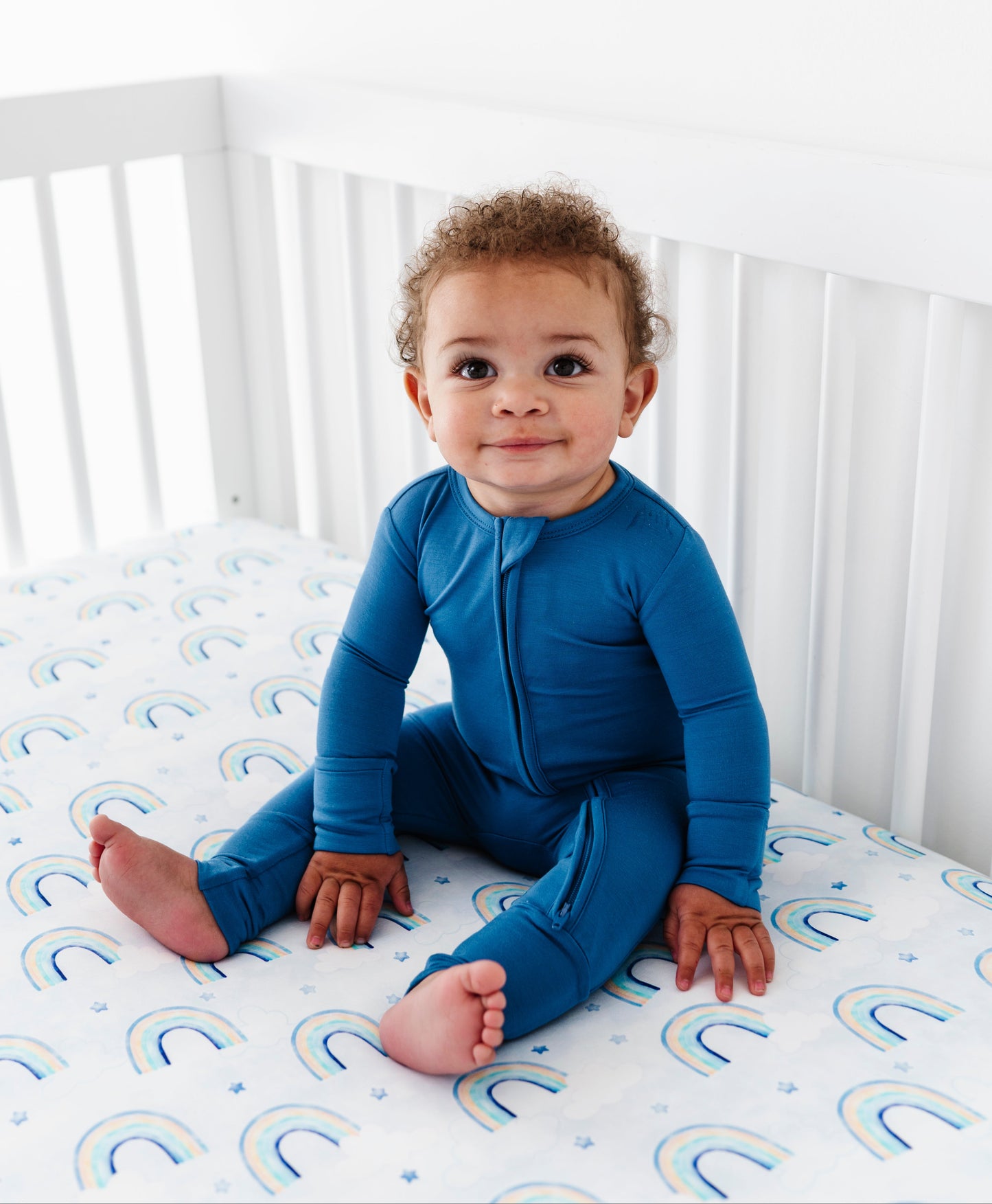 Child wearing a blue bamboo romper sitting on a crib with a mattress featuring rainbow patterns.