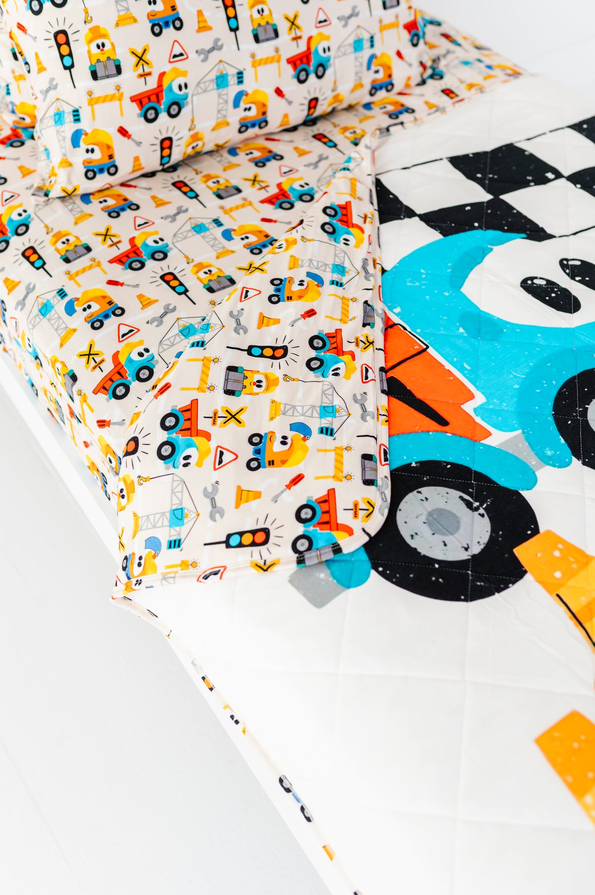 A close-up of a children's bedding set featuring a white quilted mattress cover and matching pillowcases with a colorful cartoon construction vehicle pattern, alongside a white quilted sheet with a large blue and black race car graphic.