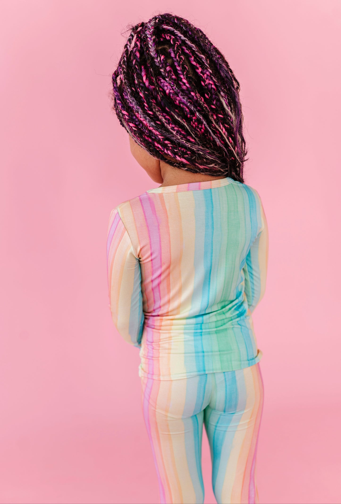 A young girl wearing bamboo rainbow striped pajamas against a pink background 