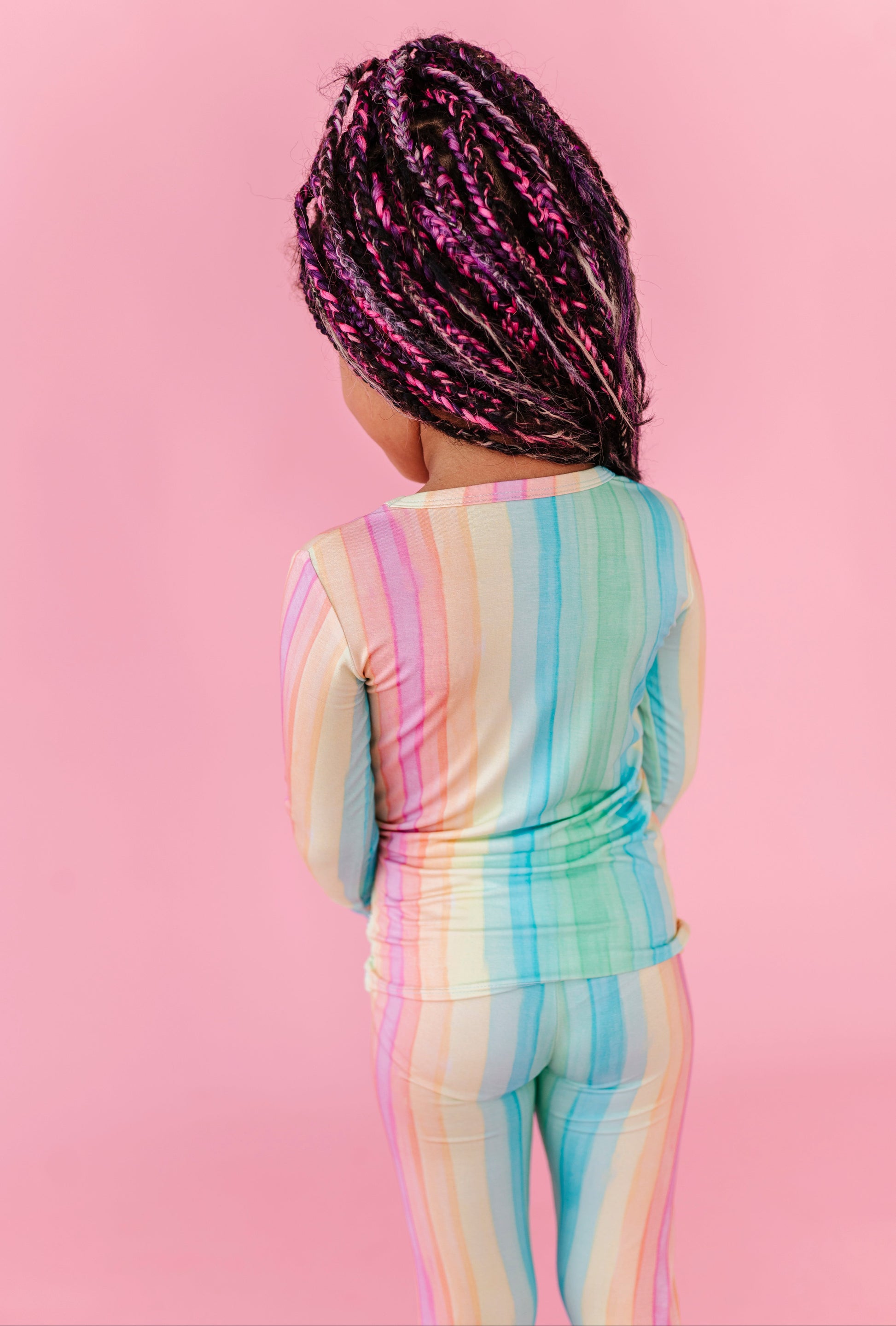 A young girl wearing bamboo rainbow striped pajamas against a pink background 