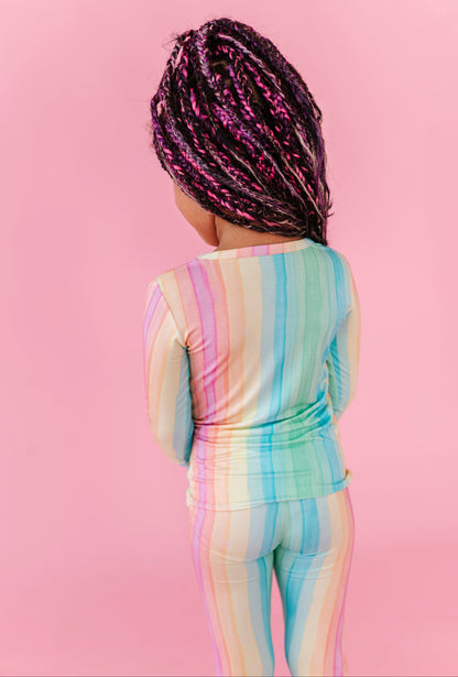 A young girl wearing bamboo rainbow striped pajamas against a pink background 