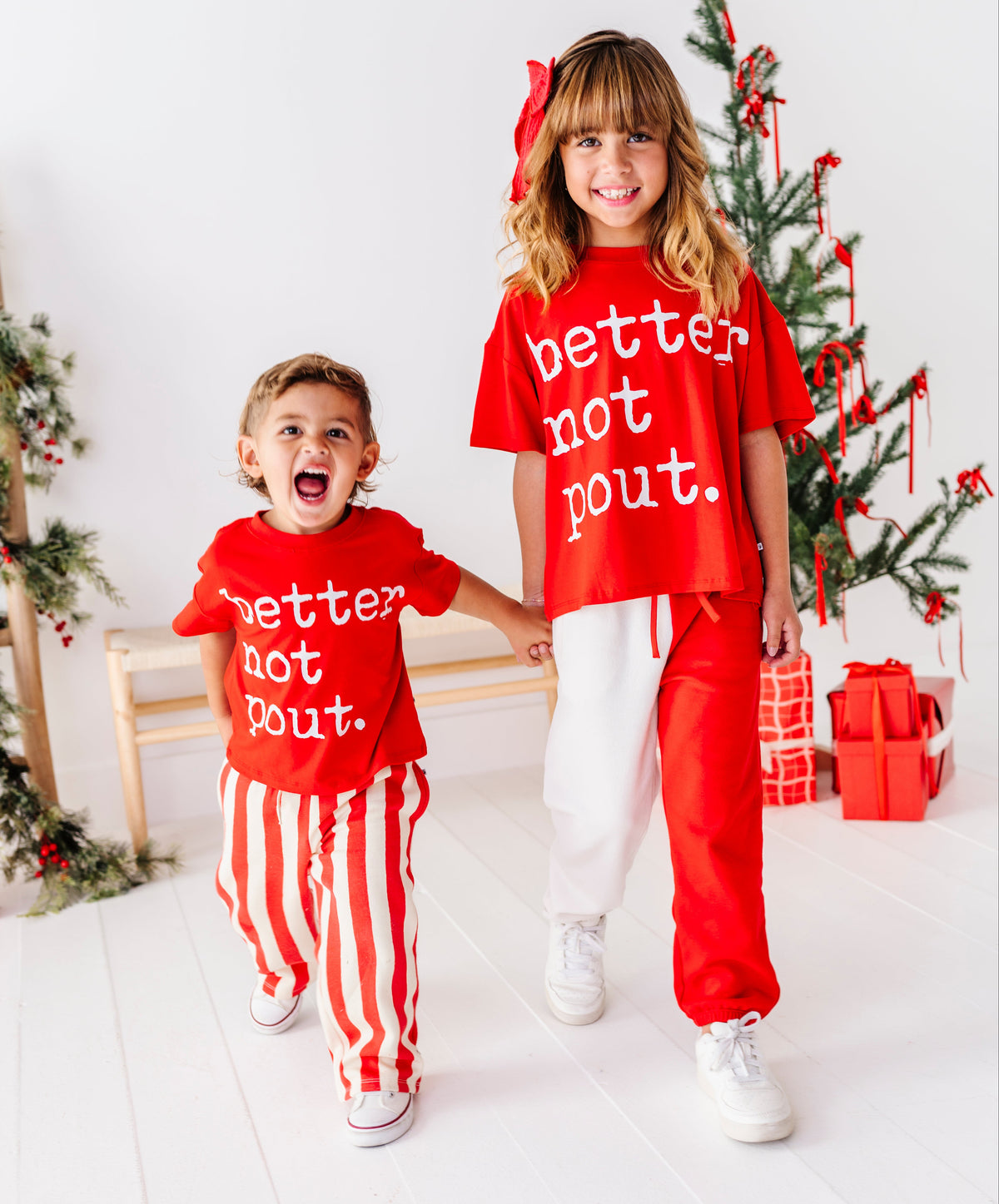 A young boy and girl wearing matching red Christmas-themed t-shirts that say 'better not bout.' in white text, walking hand-in-hand in a bright white room decorated with a Christmas tree and wrapped gifts.