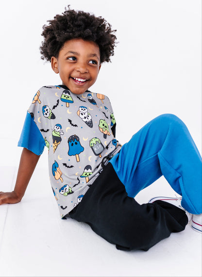 A smiling young boy with curly dark hair sits on a white studio floor wearing a gray short-sleeve t-shirt with a colorful Halloween-themed ice cream cone print, bright blue sweatpants, and white sneakers.