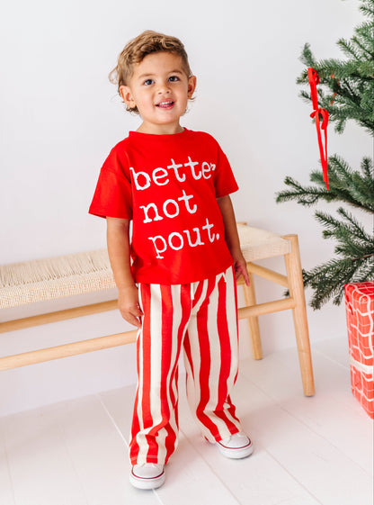 A toddler stands in a bright room wearing a red graphic T-shirt that reads 'better not bout.' and red-and-white vertically striped pants.