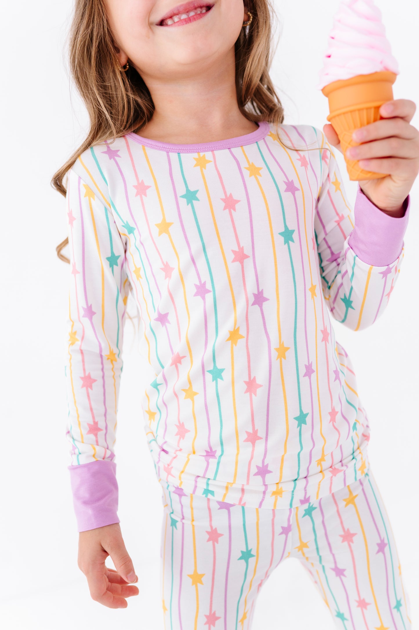 A child wearing a long-sleeve pajama top and matching pants with a pastel rainbow star and stripe pattern, holding an ice cream cone against a plain white background.