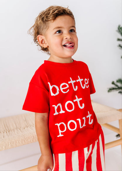 A young child wearing a bright red graphic t-shirt with the text 'bette not pout' in white, paired with red and white striped pants, standing in a brightly lit indoor setting.