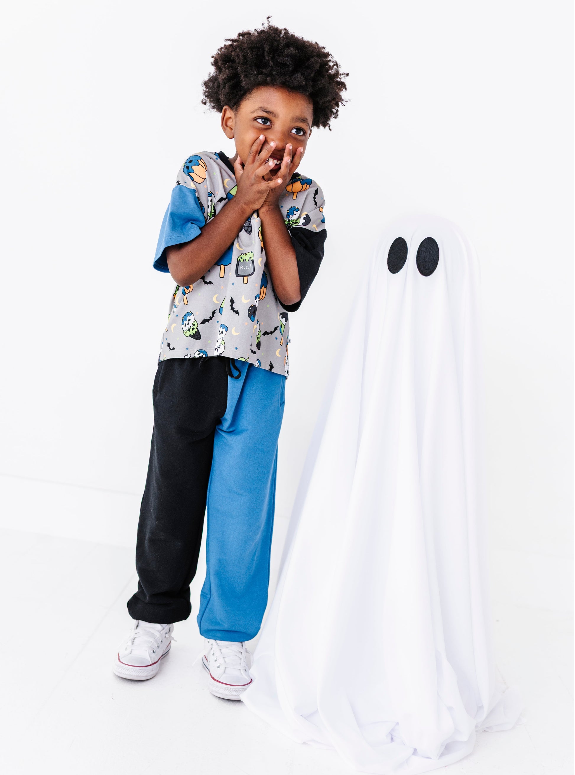 A young boy wearing a split-color graphic t-shirt, black sweatpants, and blue sweatpants stands next to a white ghost costume with black eyes on a seamless white background.