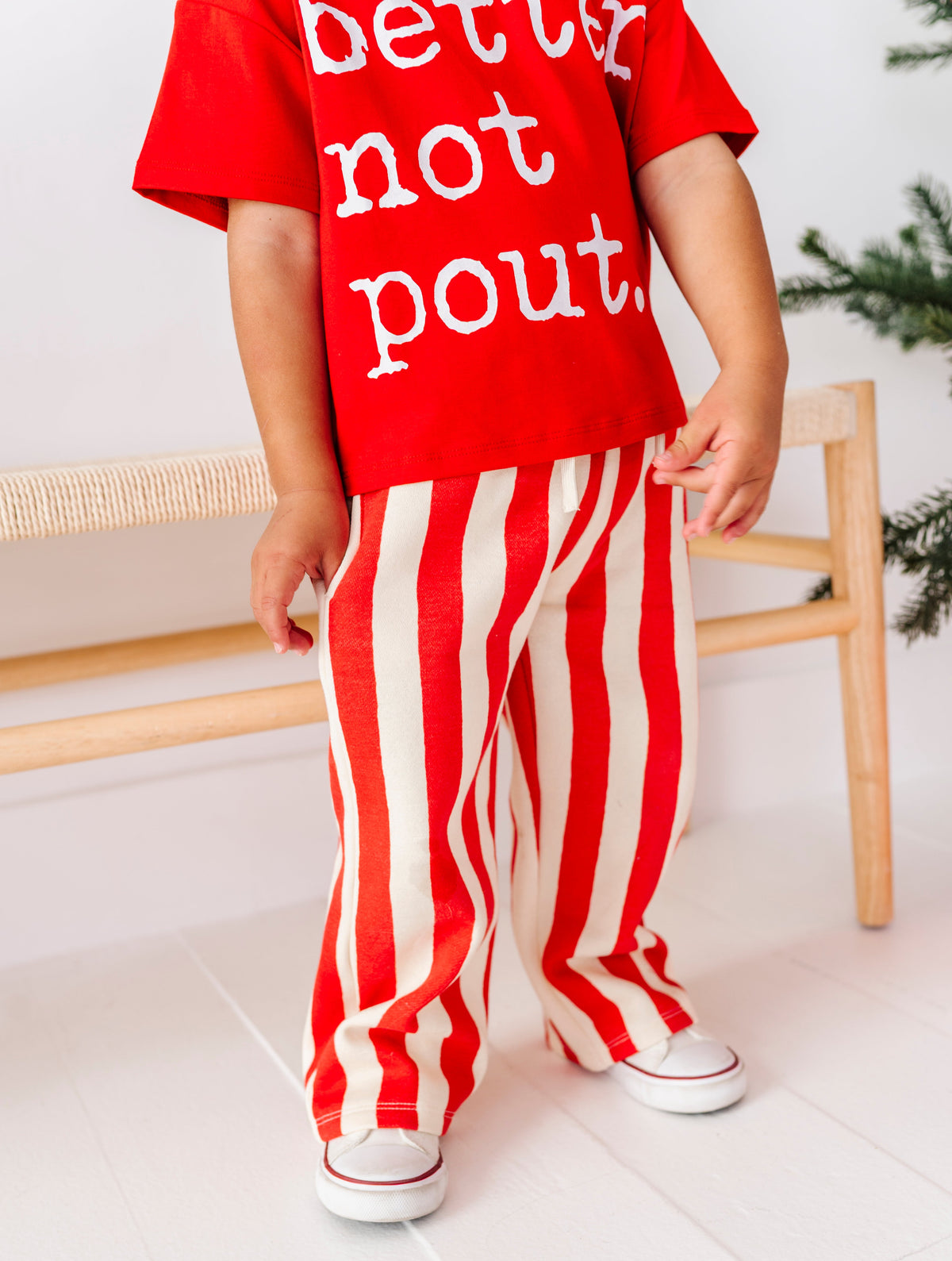 A child wearing a red short-sleeve t-shirt with white text reading 'better not pout.' and red-and-white vertically striped pants, standing indoors near a wooden bench.