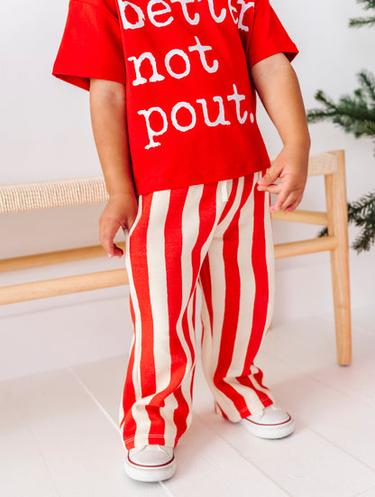 A child wearing a red short-sleeve t-shirt with white text reading 'better not pout.' and red-and-white vertically striped pants, standing indoors near a wooden bench.