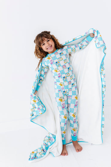 Child wrapped in a colorful 44 Cats blanket with cartoon cats like Lampo and Milady wearing matching two-piece pajamas on a white background 