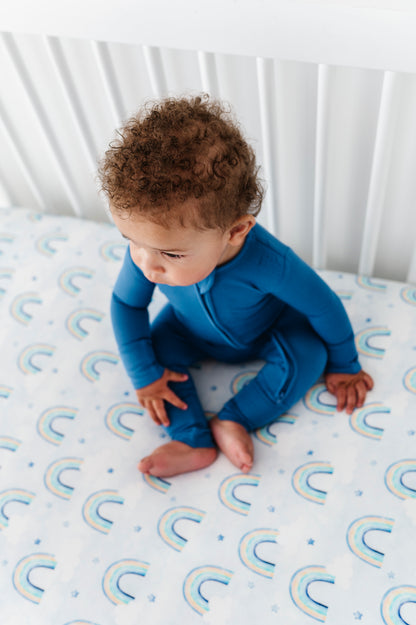 Baby in a blue bamboo convertible romper sitting in a crib with a rainbow-patterned sheet.