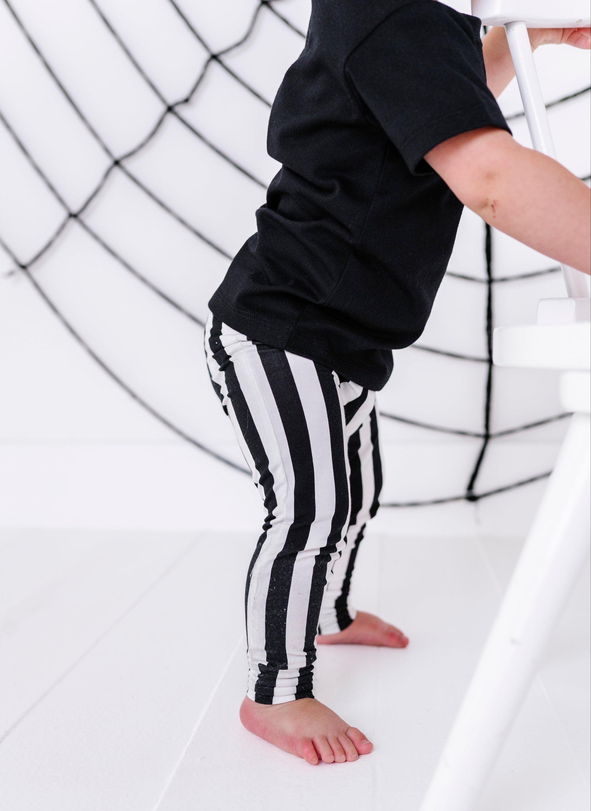 A child wearing black and white vertically striped leggings and a black t-shirt, standing barefoot on a white floor in front of a white webbed climbing wall.