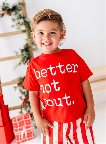 A young child wearing a bright red t-shirt with the white text 'better not bout.' and red-and-white striped pants, standing in a holiday-themed room with a ladder shelf and wrapped gifts in the background.