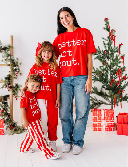 A woman and two children wearing matching red Christmas-themed apparel with white text, standing in a bright holiday-themed room with a decorated tree, wrapped gifts, and a wooden ladder.