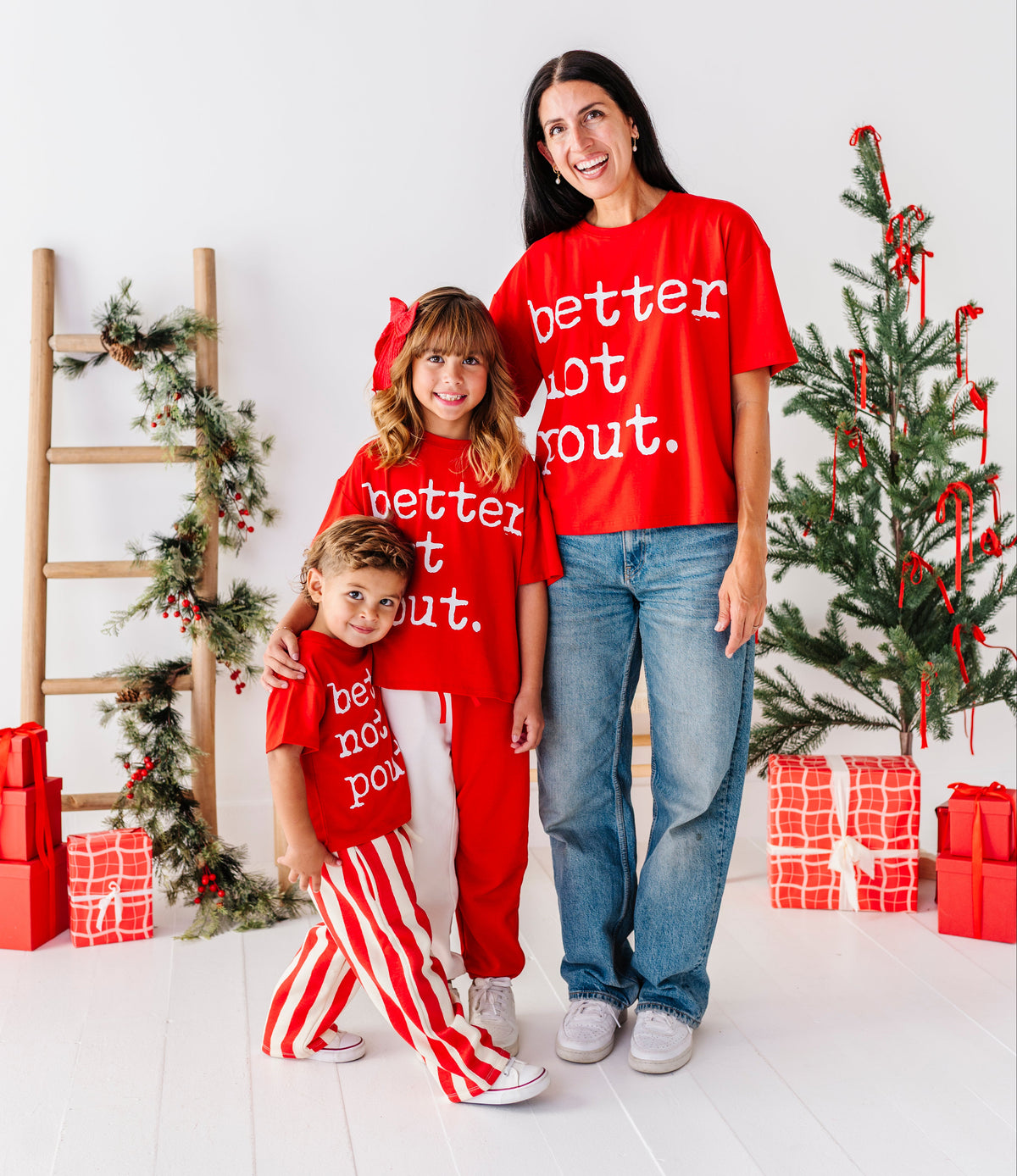 A woman and two children wearing matching red Christmas-themed apparel with white text, standing in a bright holiday-themed room with a decorated tree, wrapped gifts, and a wooden ladder.