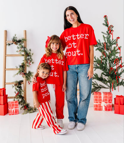 A woman and two children wearing matching red Christmas-themed apparel with white text, standing in a bright holiday-themed room with a decorated tree, wrapped gifts, and a wooden ladder.