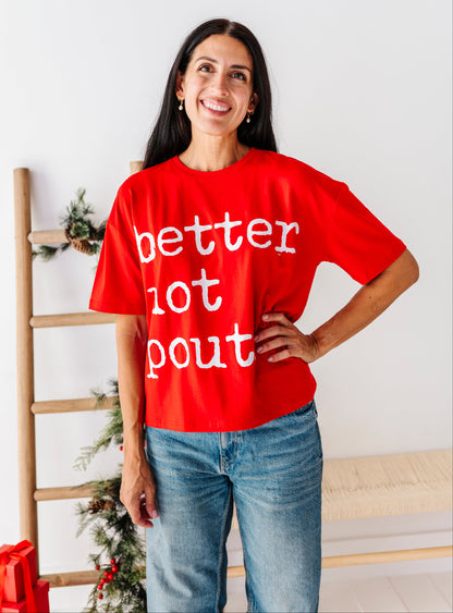 A woman wearing a bright red t-shirt with the phrase 'better not pout' printed in white lowercase letters, paired with blue jeans, standing in a brightly lit indoor setting.