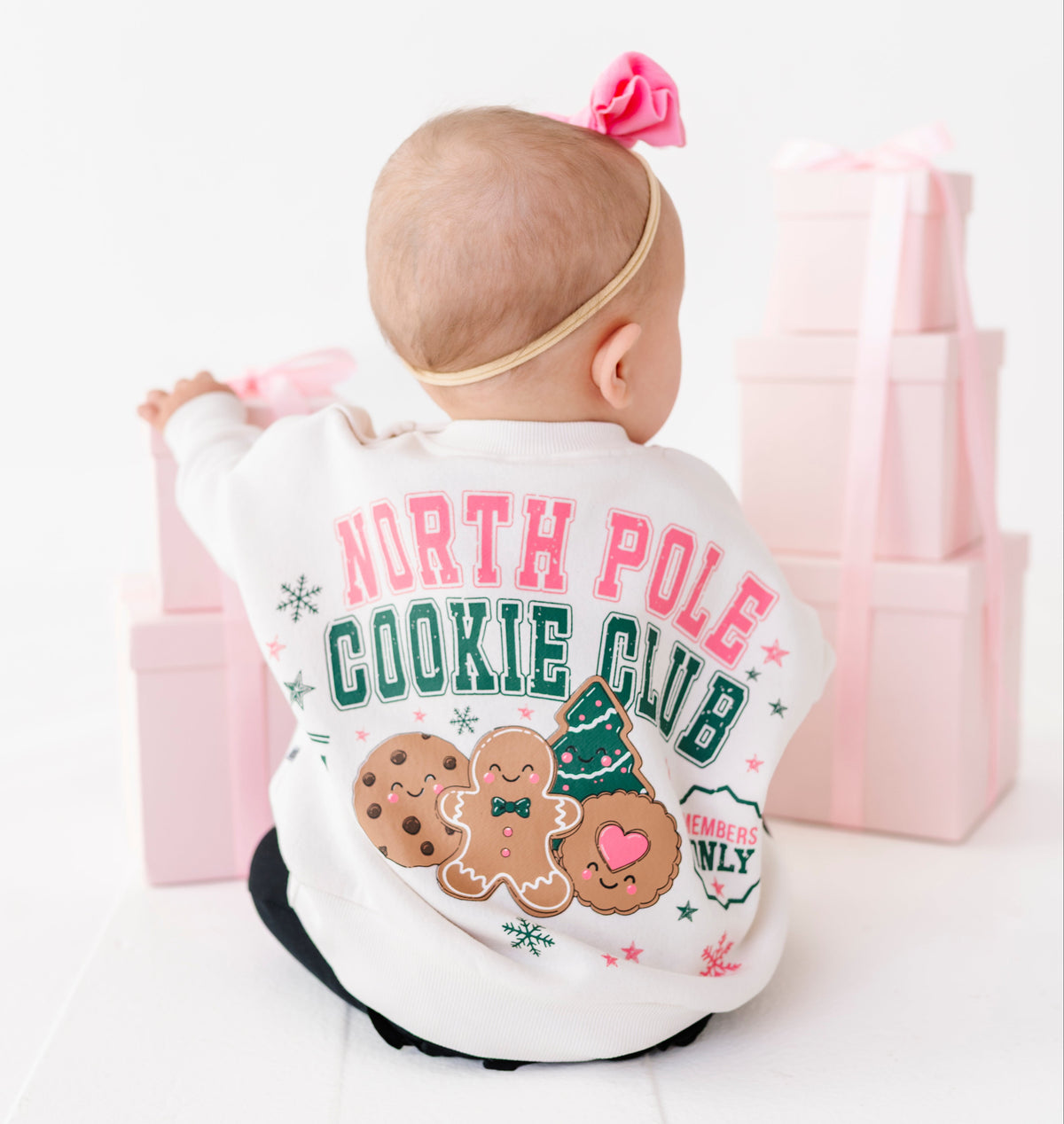 A baby wearing a white 'North Pole Cookie Club' crewneck, seen from behind, sitting on a white floor with pink gift boxes in the background.