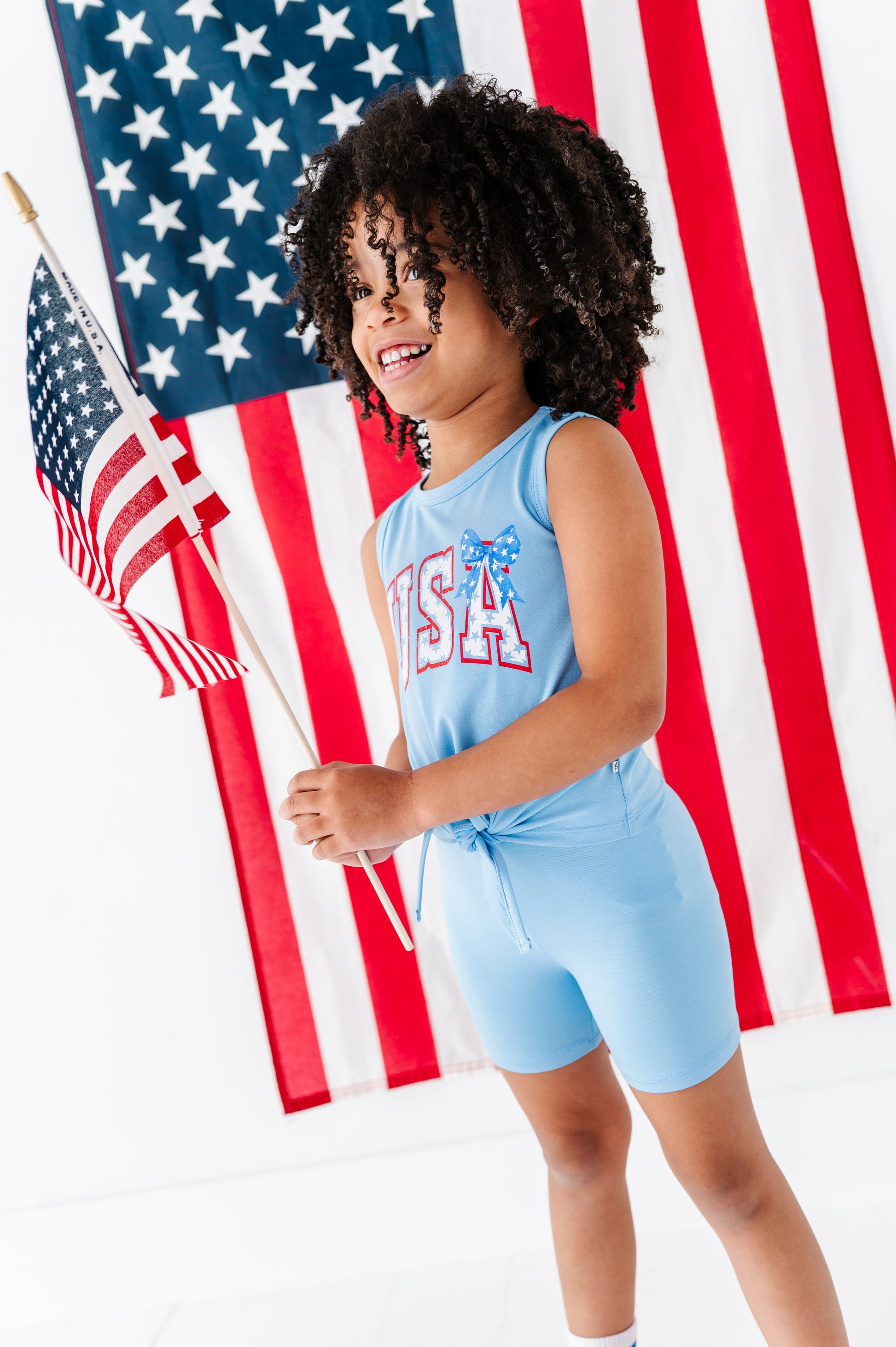 Child wearing a blue 'USA' tie-front tank and blue bamboo biker shorts in front of an American flag