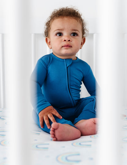 Baby in a blue bamboo zip romper sitting in a crib with a white background