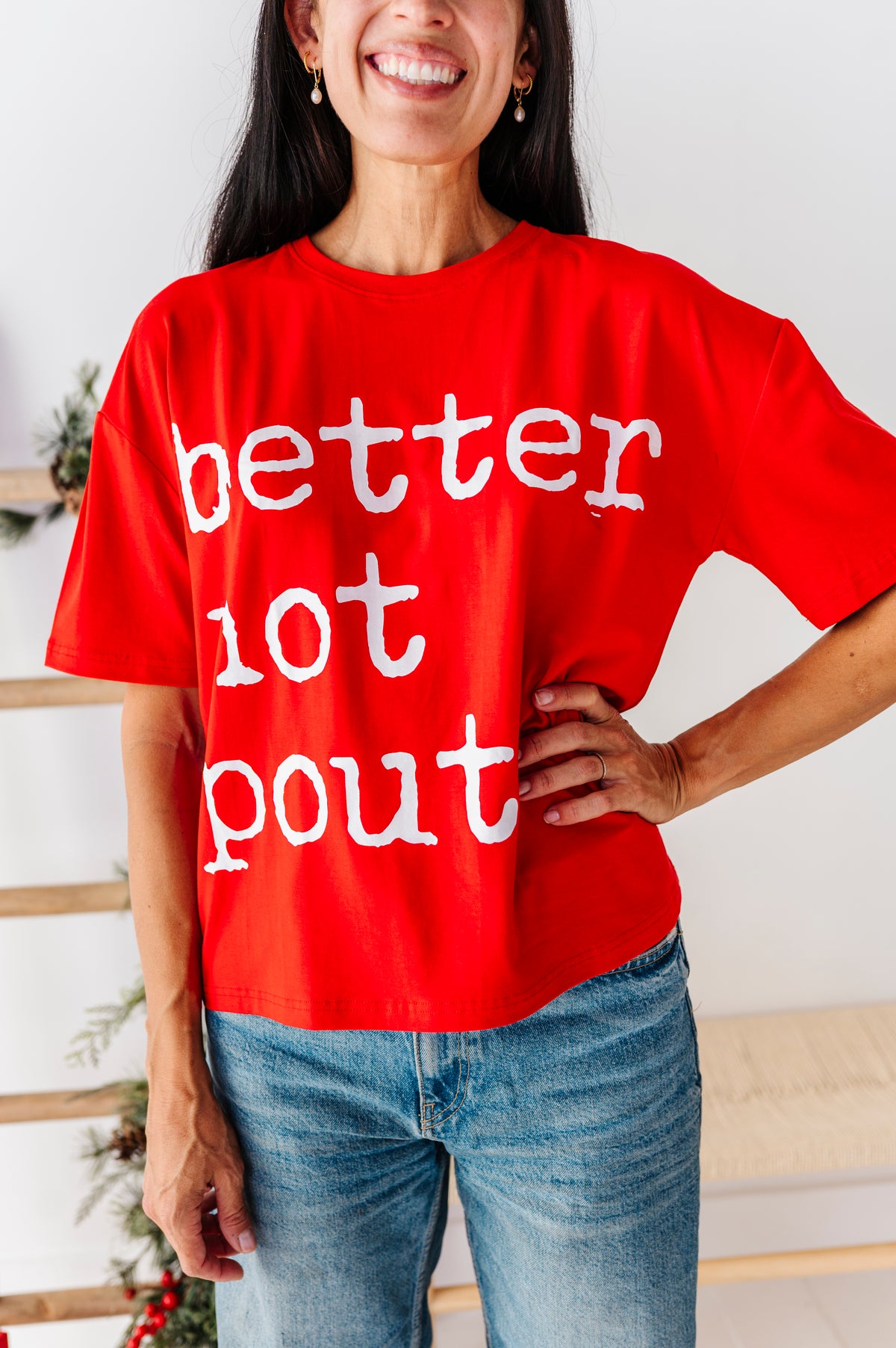 A woman wearing a bright red oversized graphic t-shirt with the text 'better not pout' in white distressed lettering, paired with blue jeans.
