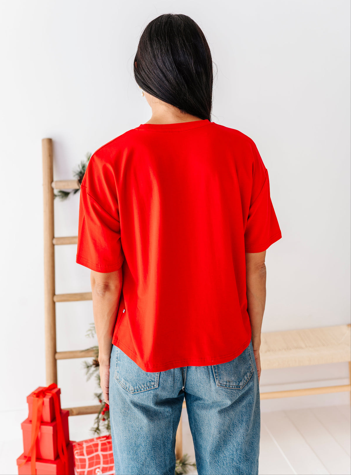 A person viewed from behind wearing a bright red short-sleeve t-shirt and blue jeans in a minimalist indoor setting with a wooden ladder and bench.