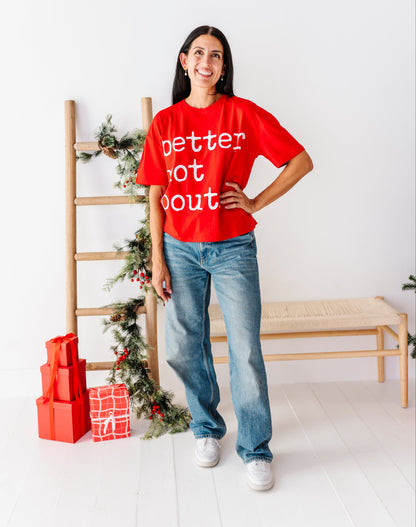 A woman stands in a bright white room wearing a red graphic t-shirt with the text 'better not out' in white, paired with blue jeans and white sneakers. A wooden bench, a ladder decorated with green garland, and red wrapped gift boxes are visible in the background.