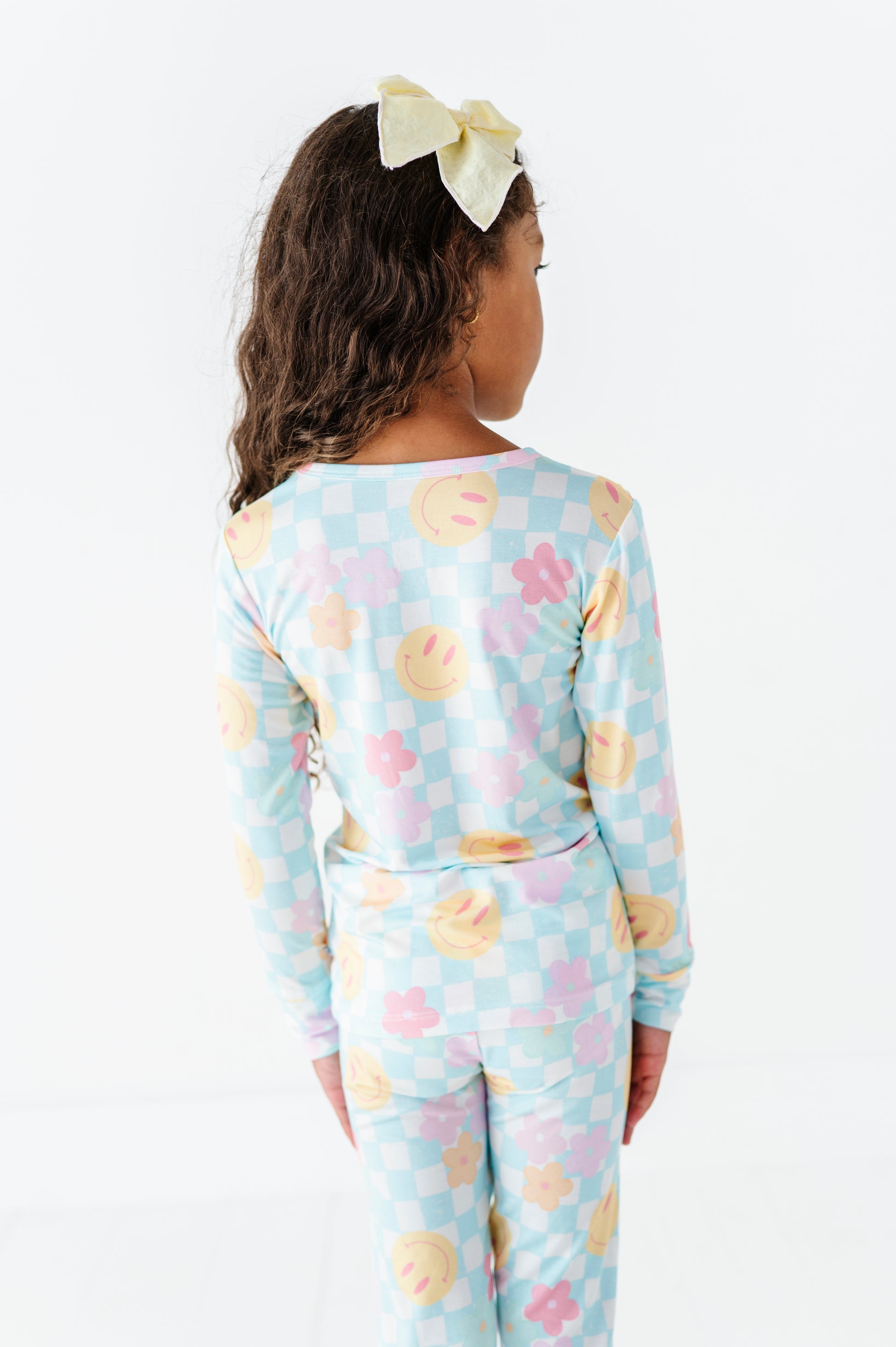 A child model shown from the back wearing a light blue long-sleeve pajama set with a pastel checkerboard pattern and pink, yellow, and orange smiley face motifs. The model has dark brown curly hair and wears a pale yellow bow headband against a clean white background.