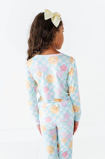 A child model shown from the back wearing a light blue long-sleeve pajama set with a pastel checkerboard pattern and pink, yellow, and orange smiley face motifs. The model has dark brown curly hair and wears a pale yellow bow headband against a clean white background.