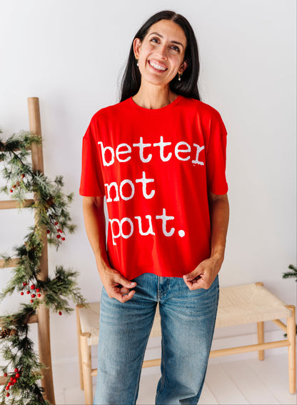 A woman wearing a bright red graphic t-shirt with the white text 'better not bout.' and blue jeans, standing in a brightly lit room with a wooden bench and holiday greenery in the background.