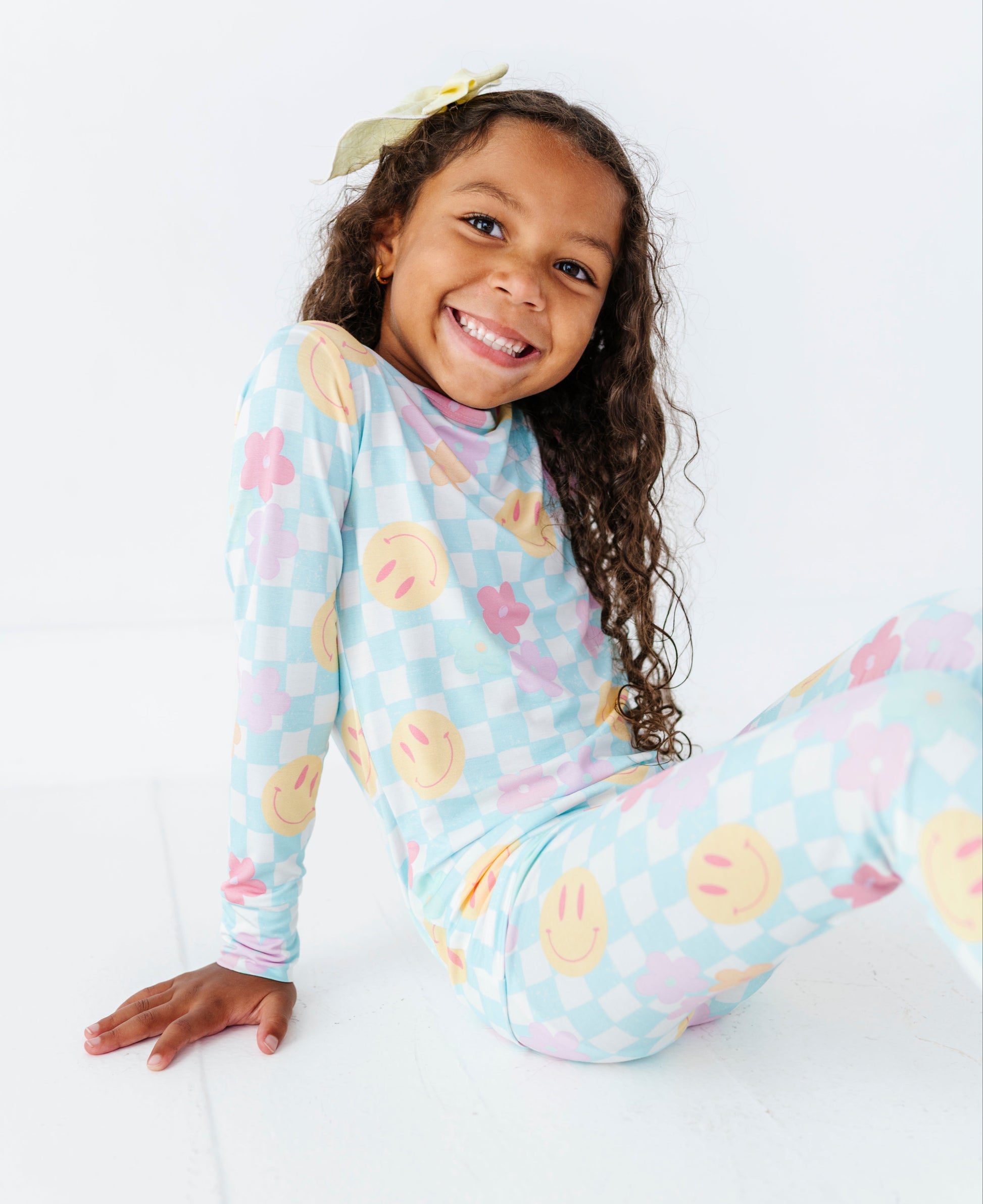 A young girl with curly brown hair wearing a light blue long-sleeve pajama set with a pastel checkerboard pattern and yellow smiley face motifs, sitting on a white floor against a white background.
