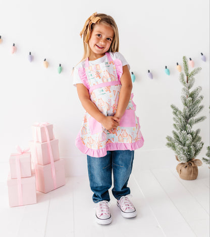 A young girl stands smiling in a bright white room, wearing a pink apron with a colorful floral print over a white t-shirt and blue jeans. Pink gift boxes are stacked on the left, and a small potted tree is on the right.