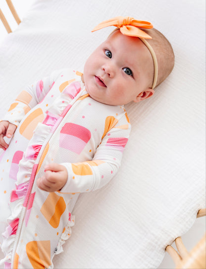 A baby lying on a white muslin mattress wearing a white long-sleeve sleep sack with pink and orange geometric cupcake patterns, along with an orange bow headband.
