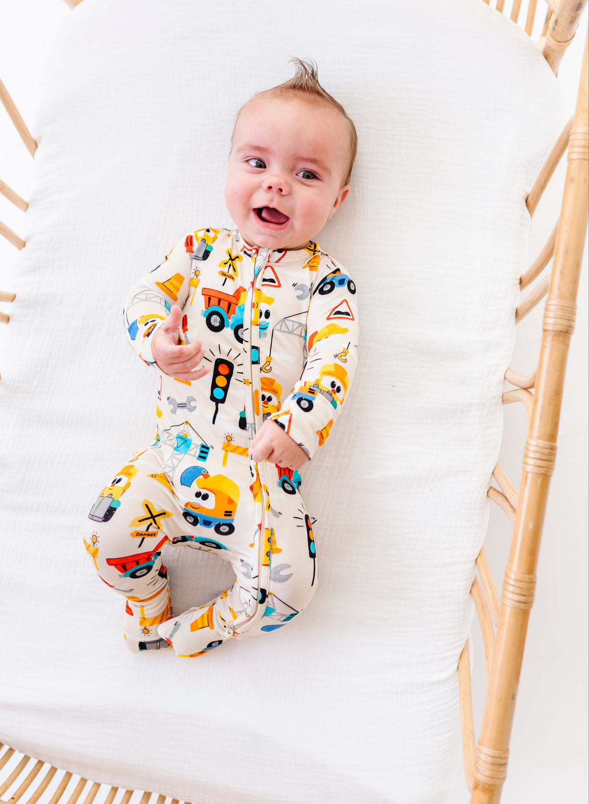 A baby wearing a colorful truck-print footed sleeper lies on a white mattress inside a light wood rattan bassinet.