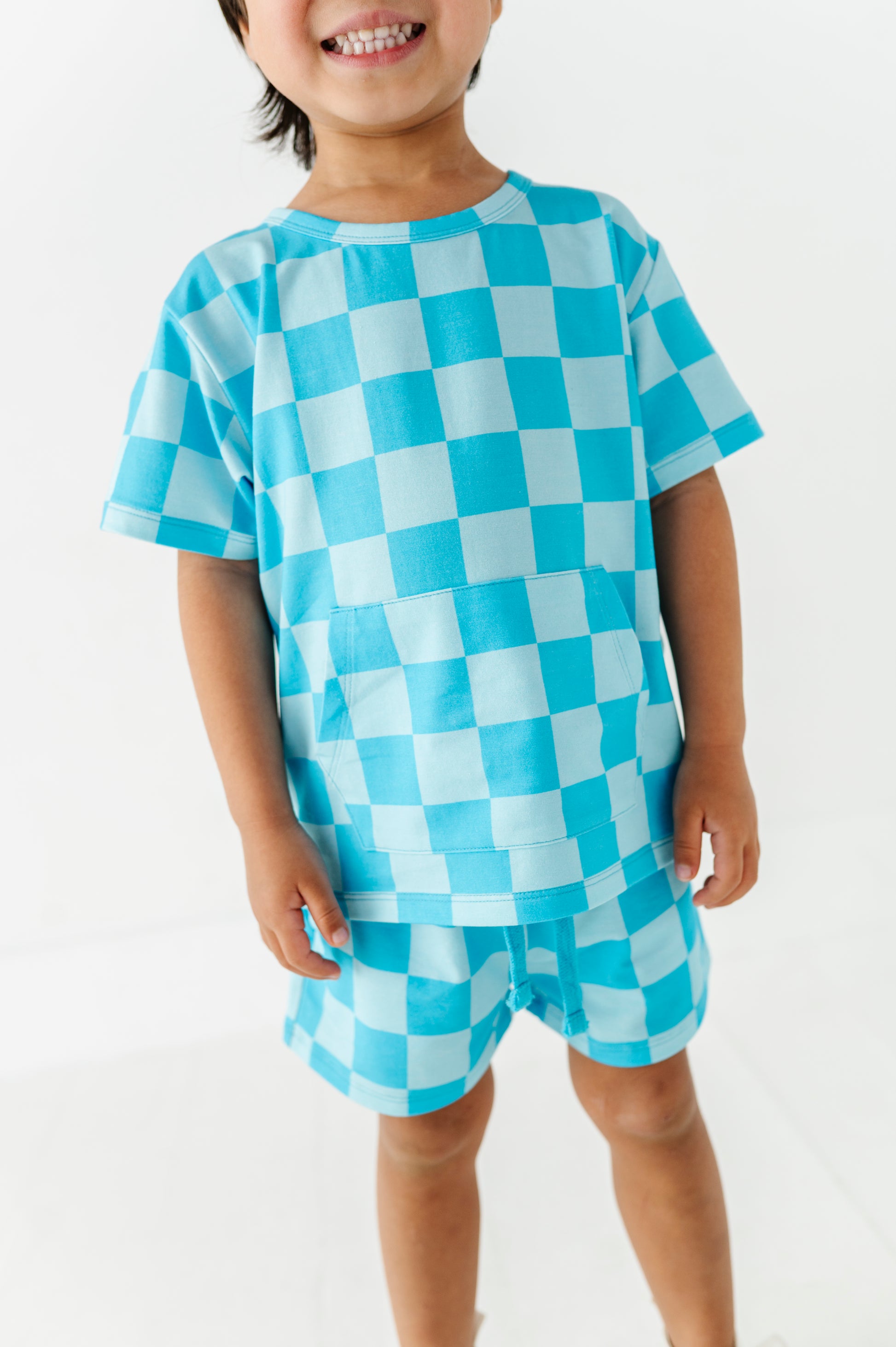 A smiling young child wearing a bright blue  checkerboard short-sleeve t-shirt and matching shorts, standing against a plain white background.