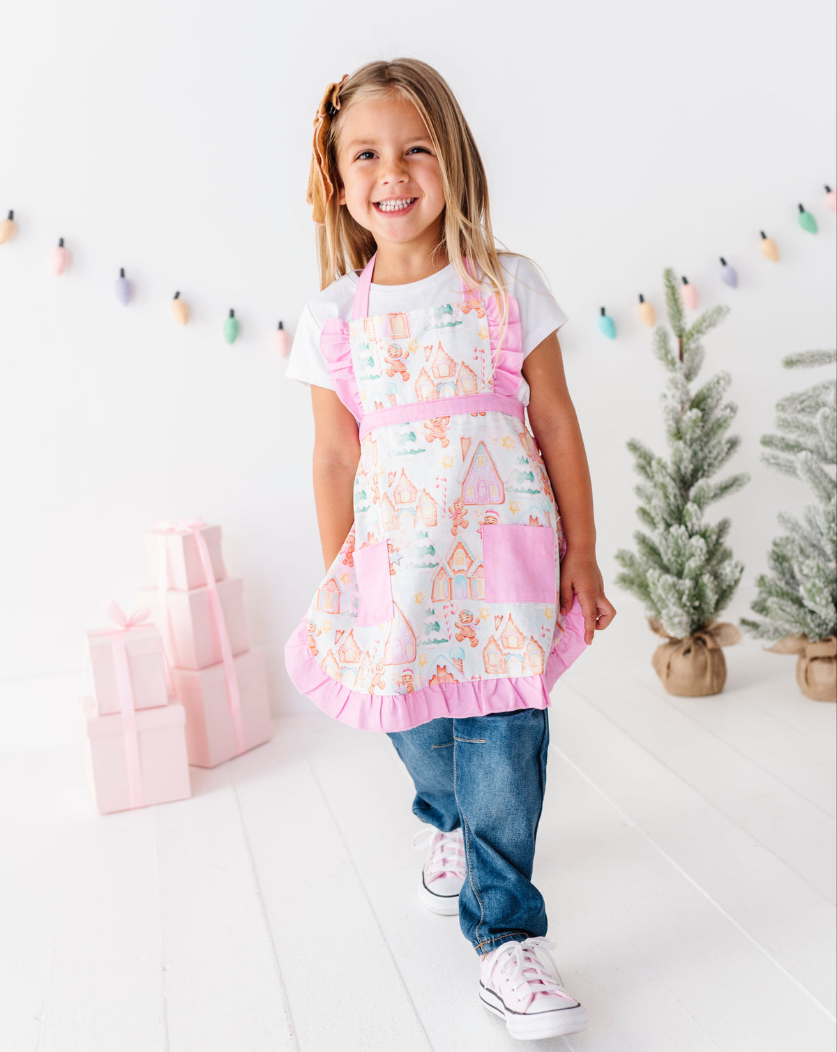 A young girl smiling and walking forward while wearing a pink and white apron with a gingerbread house pattern, blue jeans, and white sneakers in a bright, white studio decorated with gift boxes and small trees.