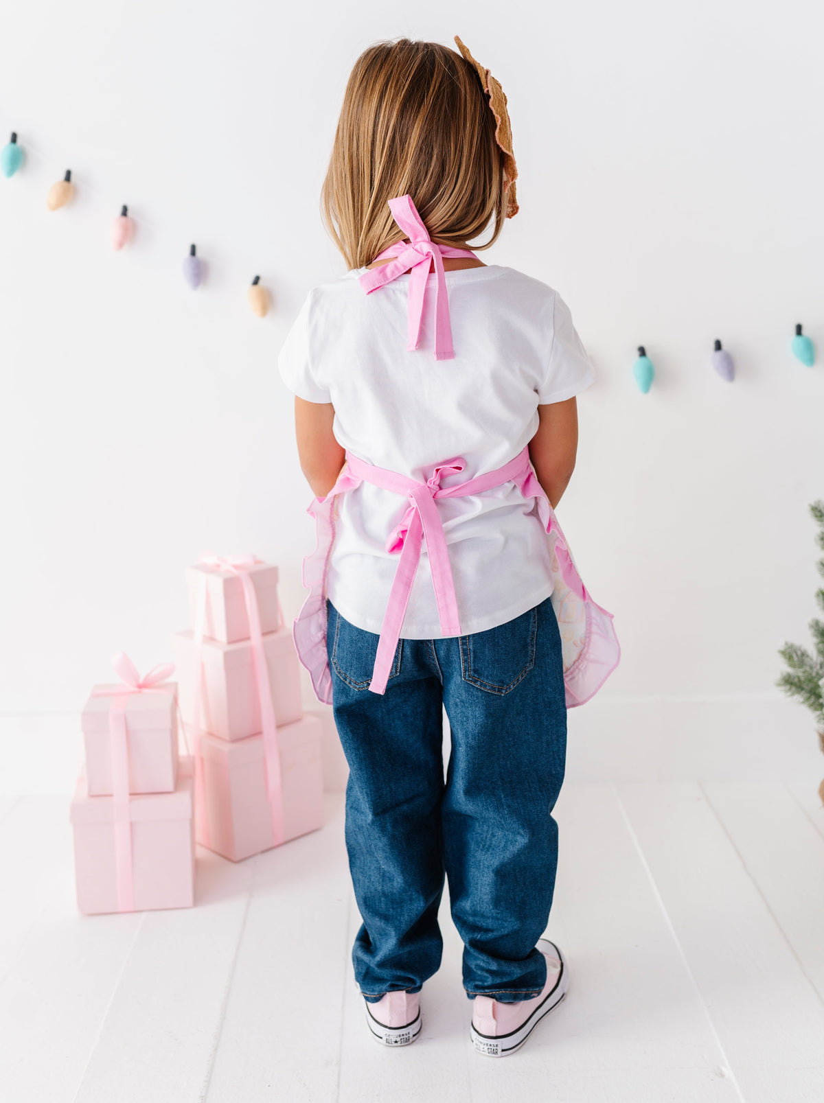 A young child viewed from behind wearing a white t-shirt, blue jeans, and a pink apron tied at the waist, standing in a bright white room with pastel gift boxes and a string of colorful hanging lights.
