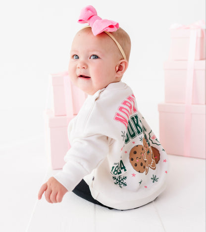A baby wearing a long-sleeve Christmas crewneck with a cookie graphic and 'NORTH POLE COOKIE CLUB' text, sitting on a white floor with pink gift boxes in the background.
