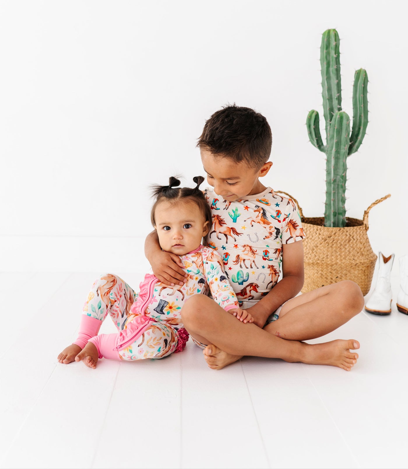 Two children in pajamas sitting on a white floor with a cactus plant in the background.