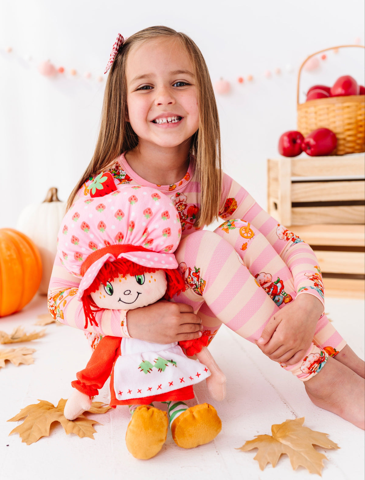 A young girl smiling while wearing a matching pink pajama set with strawberry and striped patterns, sitting on a white floor and holding a Raggedy Ann doll. The scene is decorated with autumn leaves, pumpkins, and a basket of red apples.