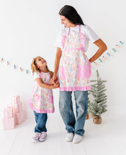 A woman and a young girl wearing matching pink and white aprons with a gingerbread-themed print, standing together in a bright, white studio decorated with a small Christmas tree and pastel gift boxes.