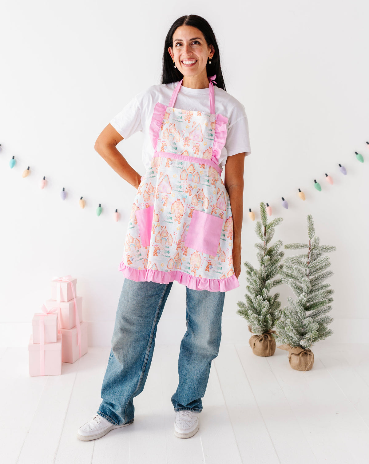 A woman stands in a bright white room wearing a pink apron with a colorful geometric pattern, a white t-shirt, and blue jeans. The apron has pink ruffled trim and pockets.