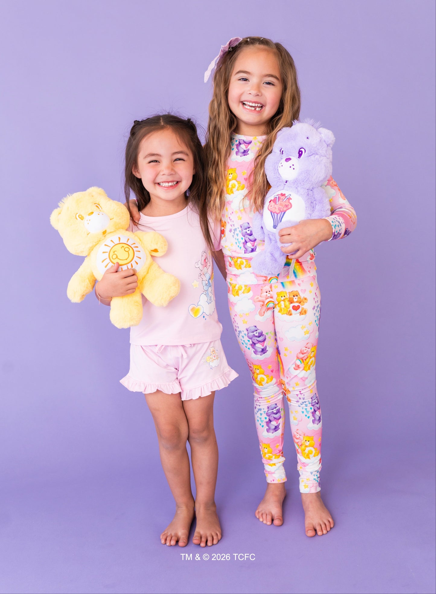 Two children in pajamas holding Care Bears™ plushies against a purple background