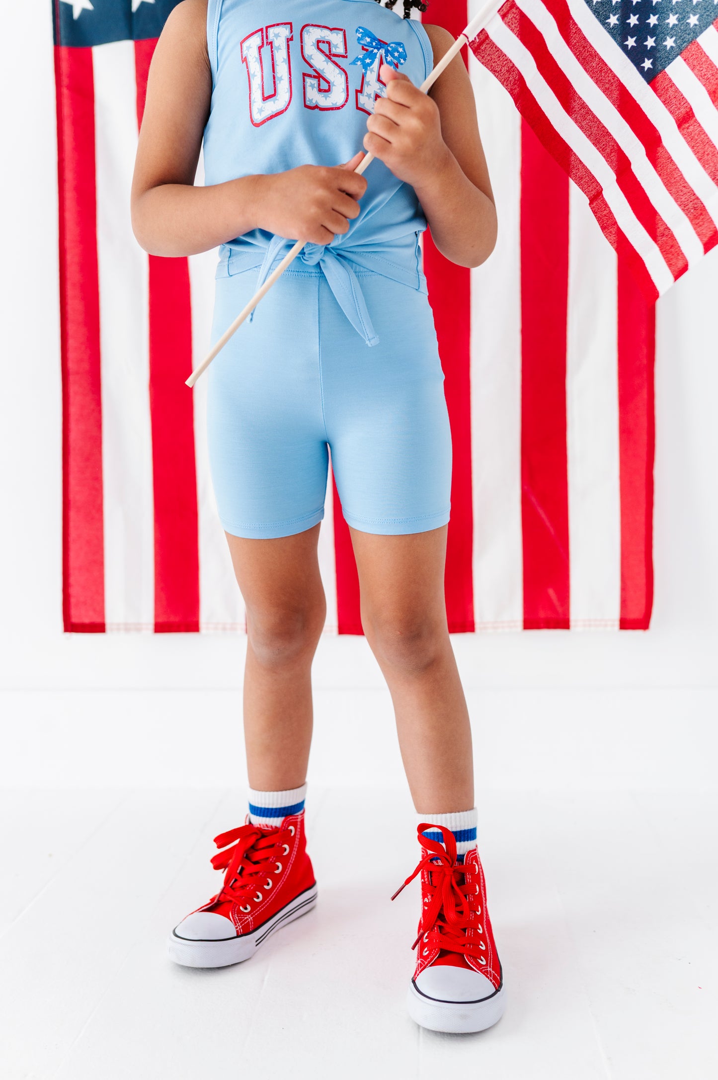 Girl wearing a light blue tank with 'USA' print, red high-top sneakers, and holding an American flag.
