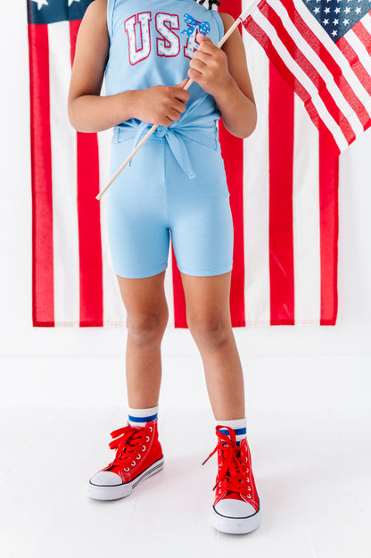 Girl wearing a light blue tank with 'USA' print, red high-top sneakers, and holding an American flag.